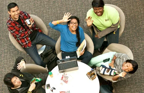 Overhead view of five students sitting around a circular table with a laptop, snacks, and drinks, engaged in reading, using phones, and interacting.