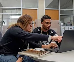 A teacher points out information on a laptop for a student sitting next to her