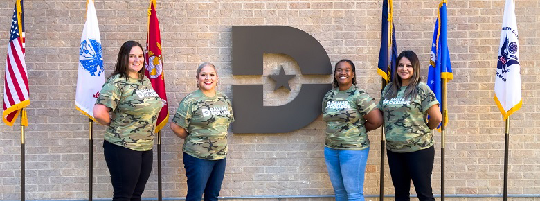 Four women veterans pose in Dallas College camouflage t-shirts near a Dallas College big D logo.