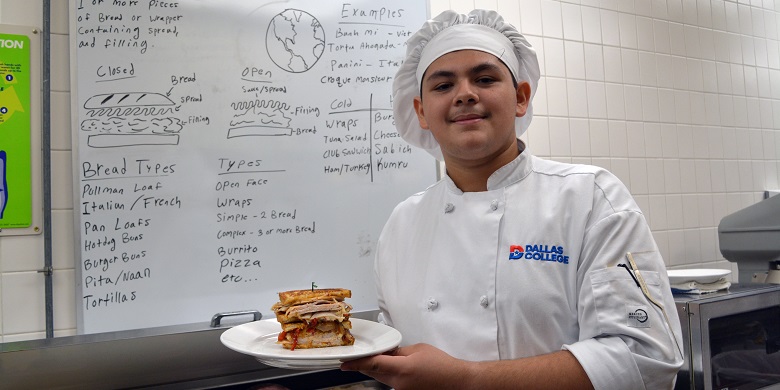 Chef Daniel Alvarenga standing in front of a whiteboard and holding plate with sandwich on it.