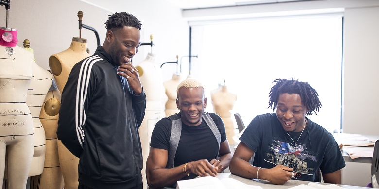 Three students gathered at a table with dress forms behind them.