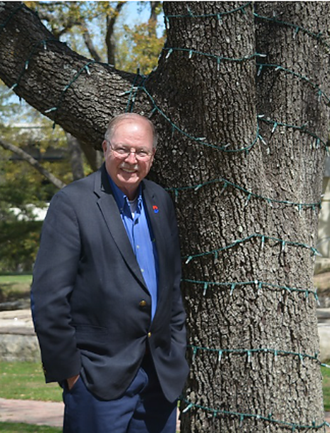Dr. Paul Benson wearing a black blazer with a Dallas college logo on the shoulder of his blazer stands next to a tree with lights( with a green wire) wrapped around it.