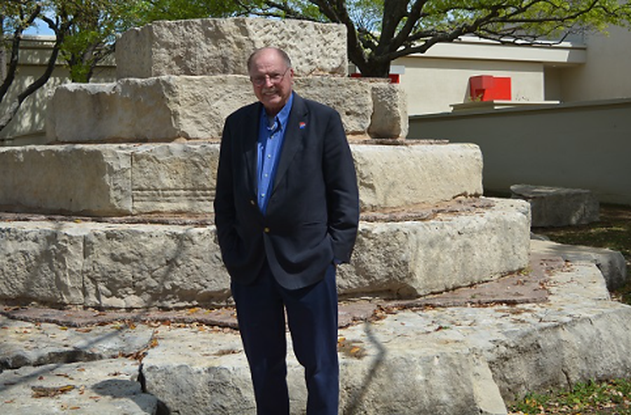 Dr. Paul Benson wearing a black blazer with a Dallas college logo on the shoulder of his blazer has his hands in his pockets and is standing in front a step-like stone structure.