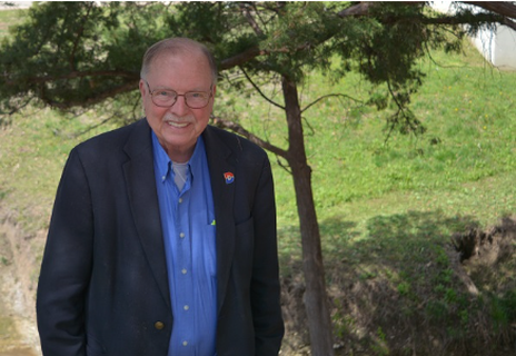Dr. Paul Benson wearing a black blazer with a Dallas college logo on the shoulder of his blazer stands in front of a tree and grass hill.