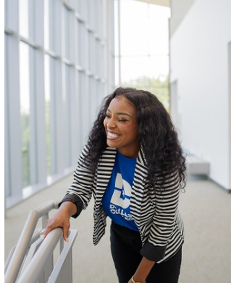 Kianna Vaughn in a blue shirt with a white Dallas College logo and a blazer is smiling while holding a railing.