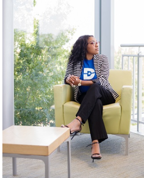 Kiana Vaughn is wearing a blazer, looking away from the camera , and sitting cross legged on a white armchair.