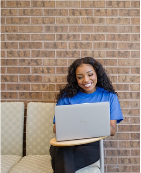 Kianna Vaughn in a blue shirt with a white Dallas College logo is smiling while working on a computer on a white and gray gingham couch.