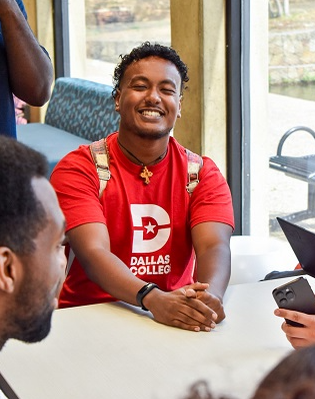 Konata Kidane wearing a red shirt with the Dallas College logo and a backpack, looks upward to the camera while sitting at a table.