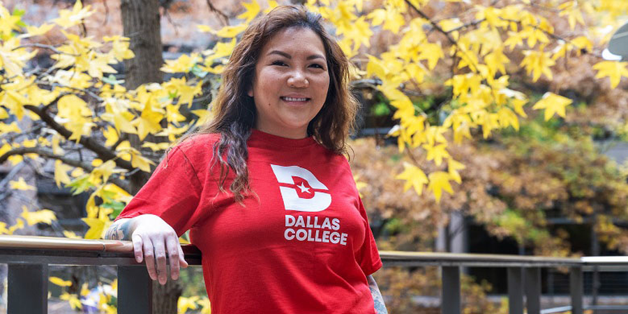 Ngoc Trinh in a red shirt with the Dallas Logo logo on it stands by a railing with autumn trees behind.