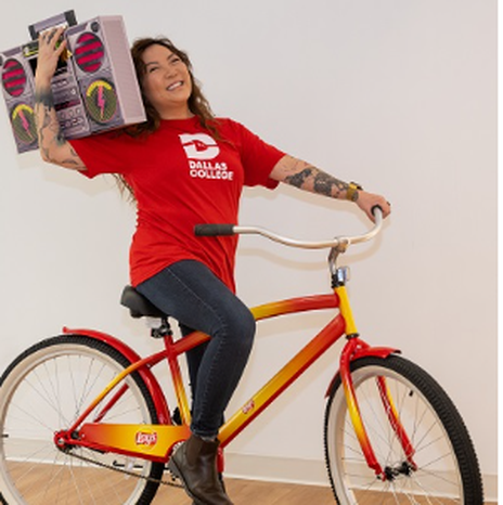 Ngoc Trinh in a red shirt with the Dallas Logo logo is on a ride bicycle holding a pretend (maybe cardboard) boombox.