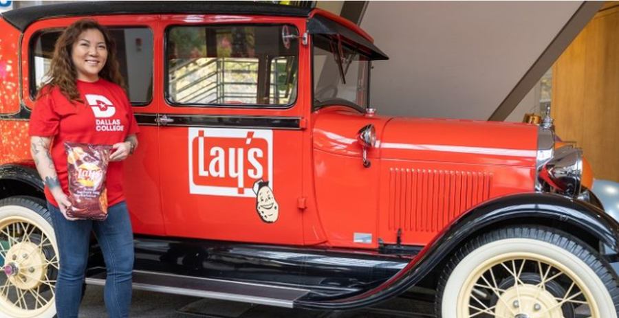Ngoc Trinh in a red shirt with the Dallas Logo logo on it holds a bag of Lay's sweet southern heat barbecue chips while standing in front of a red vehicle with the Lays logo on it.