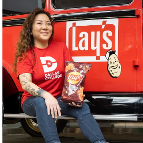 Ngoc Trinh in a red shirt with the Dallas Logo logo on it holds a bag of Lay's sweet southern heat barbecue chips while sitting in front of a red vehicle with the Lays logo on it.