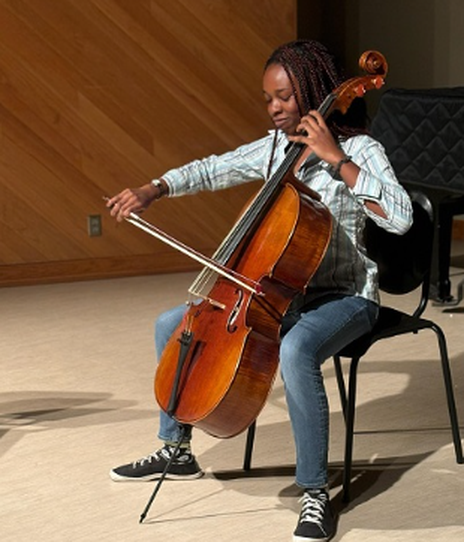 Skylar Cambell plays the Cello in front of a piano with a quilt like cover on it and a wall with a wooden design..