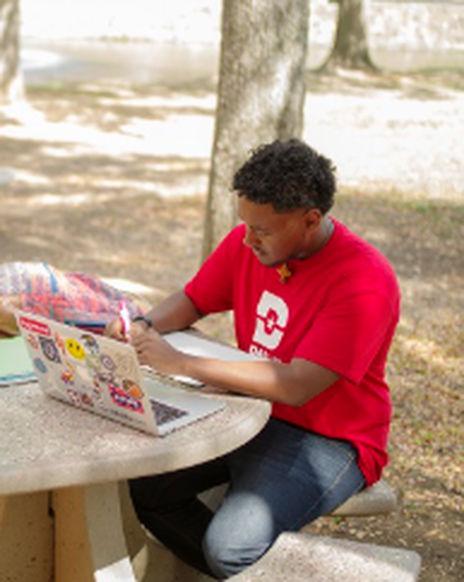 Yonata Kidane wearing a red shirt with the Dallas College logo is working outside on a picnic table using a computer, notebook, a pen..
