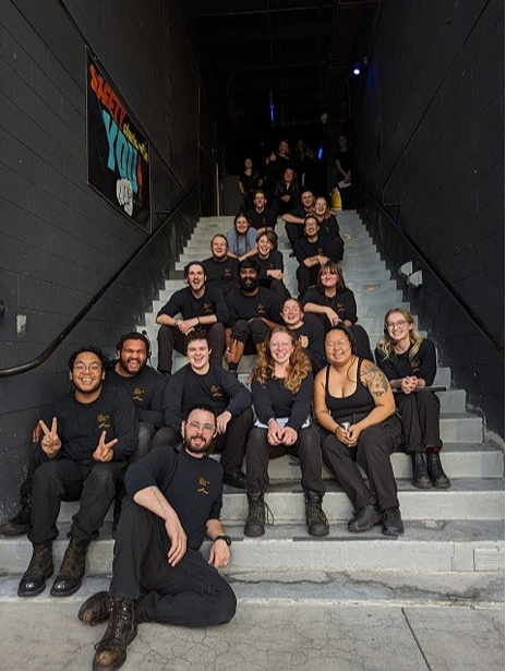 Stipe sitting with the whole crew, with everyone sitting on a set of stairs and wearing black. They all pose in different ways and smile towards the camera.