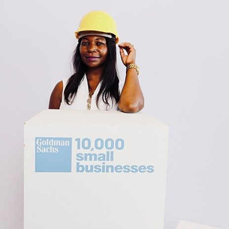 Ebbah at the Goldman Sachs 10,000 Small Businesses program, standing behind a podium with the initiative’s logo. She wears a yellow construction hat, with her left hand holding it. She smiles in front of a plain white background