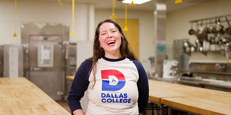Frida Hutchinson standing in a kitchen wearing a Dallas College shirt, smiling at the camera.