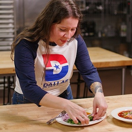 Frida Hutchinson preparing a plated dish during a culinary demonstration at Dallas College. She is wearing a Dallas-themed shirt while working in the college’s kitchen space.