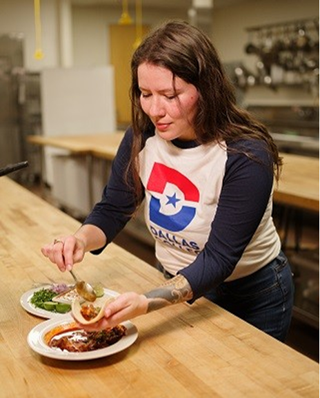 Frida Hutchinson preparing a plated dish in a professional kitchen, wearing a Dallas College T-shirt. She is using both of her hands, with one holding the base of the food, and the other to hold a spoon for filling.
