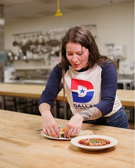 Frida Hutchinson preparing a dish in a Dallas College kitchen, wearing a shirt with the college's logo