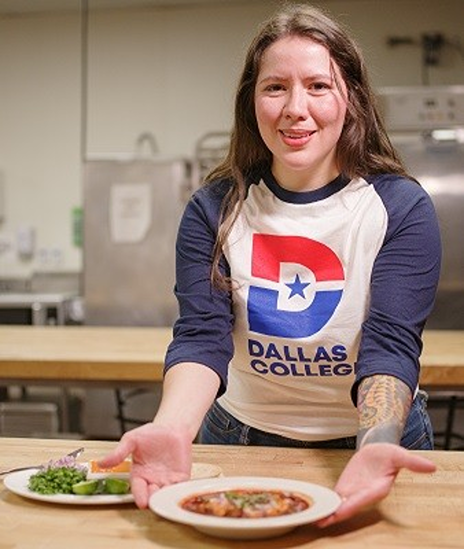 Frida Hutchinson presenting a plated meal in a kitchen setting while wearing a Dallas College T-shirt