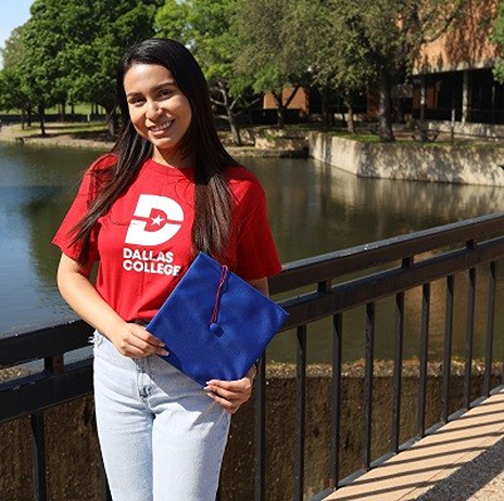 Pricila Cano Padron wears a red Dallas College shirt and holds her college cap in her hands, as she smiles towards the camera. Nature is visible in the background.