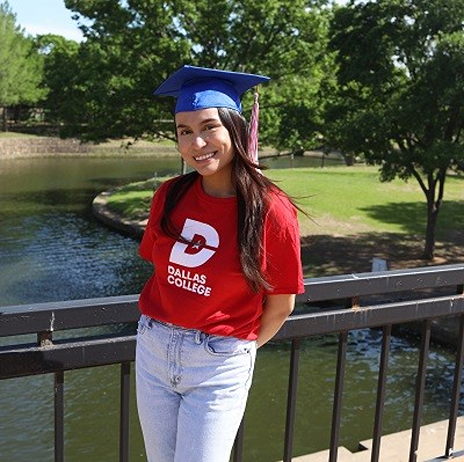 Pricila Cano Padron wears a red Dallas College shirt with her hands behind her back and wears a graduation cap. She stands on a bridge, with a fountain in the background and nature.