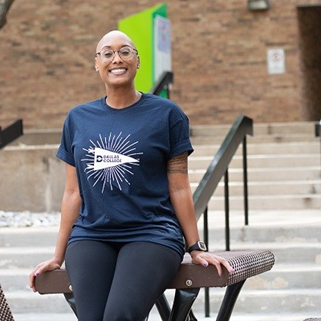 Qualonda Bennett sitting on a table, with her hands on the table and her legs crossed. She wears a Navy Dallas College shirt while smiling and facing the camera. A building of a Dallas College campus is visible in the background.