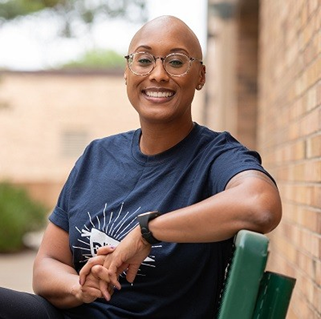 Bennett sitting at a green bench, with her fingers intertwined. She wears a navy Dallas College shirt, as she smiles towards the camera. A Dallas College campus is visible in the background.