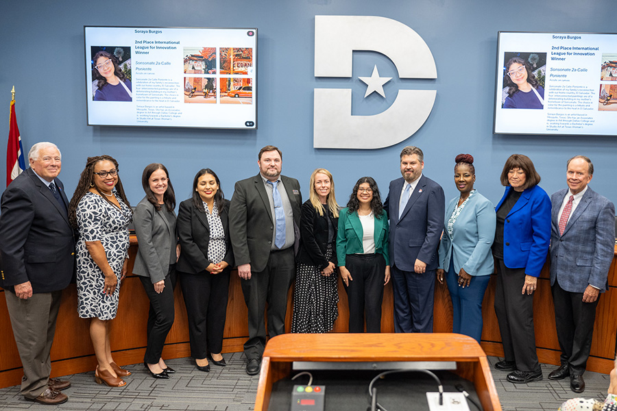 Soraya Burgos stands with Dallas College leaders and board members during a recognition ceremony honoring her success of her art. Presentation screens behind the group display images of the artwork and information about Burgos' with a Dallas College logo in the center.