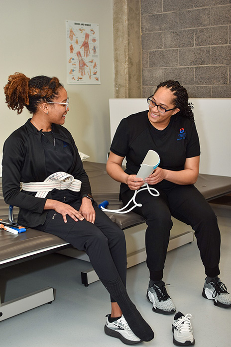 Tometra Williams wearing a black shirt with the Dallas College logo on her upper left corner of the shirt is teaching a patient who has her shoe off.