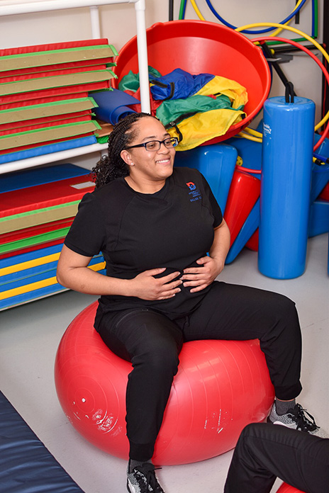 Tometra Williams wearing a black shirt with the Dallas College logo on her upper left corner is sitting on a large, red medicine ball with her hands on her stomach.
