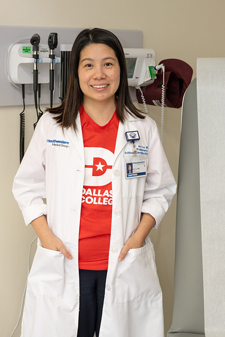 Dr. An Lu standing in front of wall-mounted medical equipment and wearing a lab coat and Dallas College t-shirt.