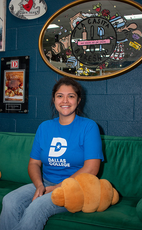 Maricsa sits on a green couch, sitting next to a croissant pillow inside her bakery, La Casita Bakeshop.