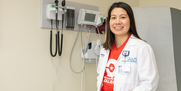 Dr. An Lu standing in front of wall-mounted medical equipment and wearing a lab coat and Dallas College t-shirt.