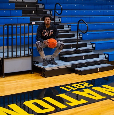 Antoine Silvers Jr sitting on the stands, holding a basketball at his knee-level, facing the camera inside a Dallas College gym.
