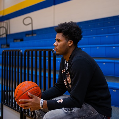 Antoine Silvers Jr sitting on the stands, holding a basketball at his knee-level, facing away from the camera as the photo is take from the side. The photo is taken inside a Dallas College gym.