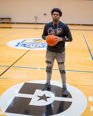 Antoine stands around the center of a basketball court on a Dallas College campus, where a Mountain view and Dallas College logo is visible. He holds a basketball with two hands around his chest as he faces the camera.