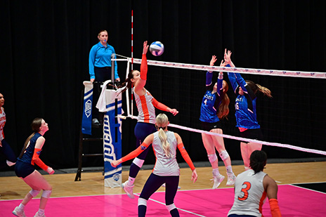 Eastfield Women's volleyball player trying to hit the ball over the net, with the opposing team trying to block it wearing blue in a Championship game. The other Eastfield players watch.
