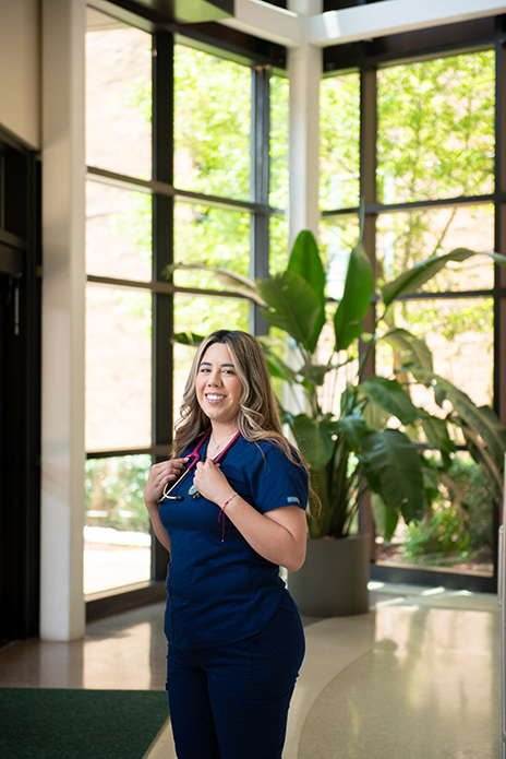 Faviola Cecena in blue scrubs wearing a stethoscope around her neck is standing in front of a wall made of windows and an indoor plant.
