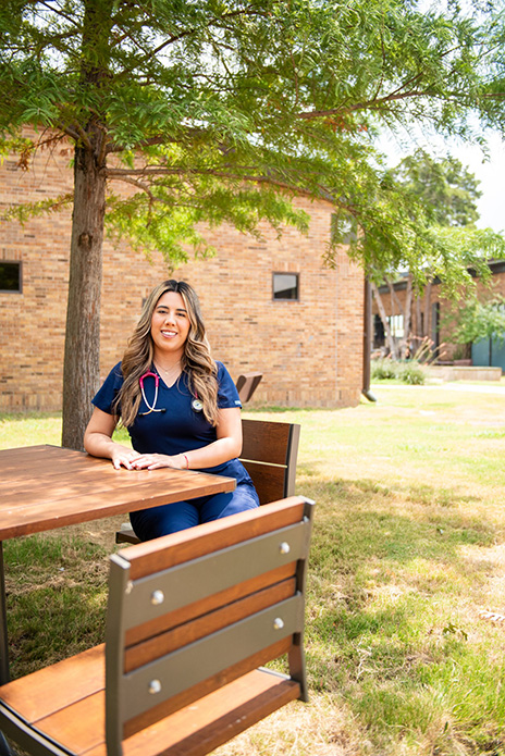 Faviola Cecena in blue scrubs wearing a stethoscope around her neck is sitting at a brown, rectangular table.