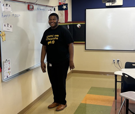 Elijah stands at the front of a classroom beside a whiteboard displaying the date and classroom instructions. A Texas flag, projection screen and student desks are visible in the classroom.
