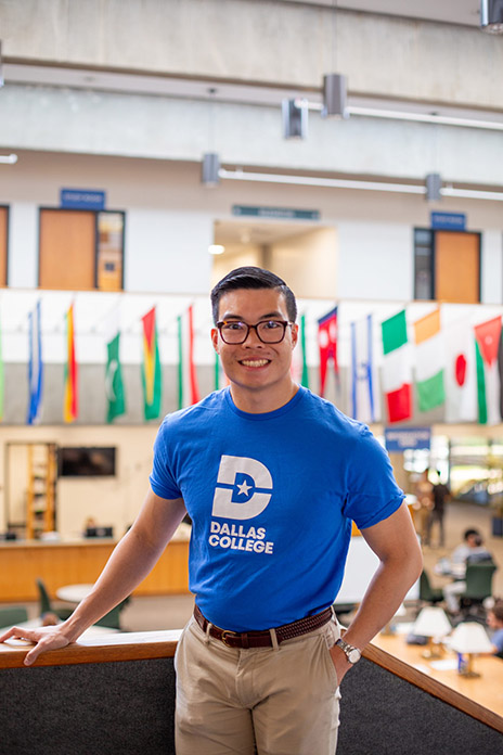 Harrison confidentally standing and smiling at the camera in a Dallas College library.