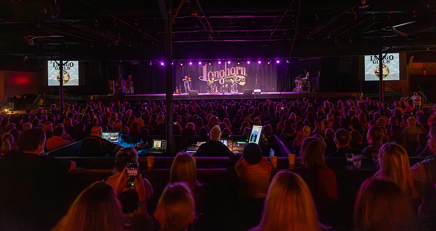 A crowd gathers inside the Longhorn Ballroom during a live concert. The audience faces a brightly lit stage, where technicians work from a control area near the center of the venue.