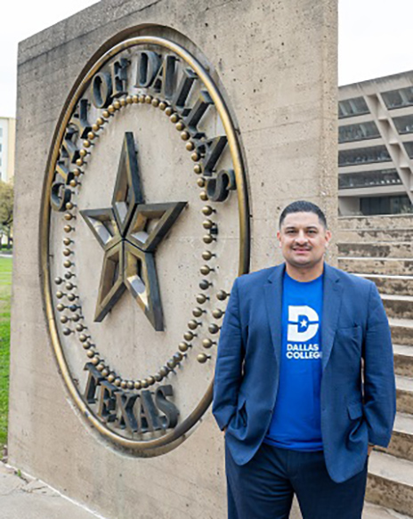 Dallas College alum Jaime Resendez stands outdoors in front of a City of Dallas sign, wearing business-casual attire and posing confidently on a walkway.