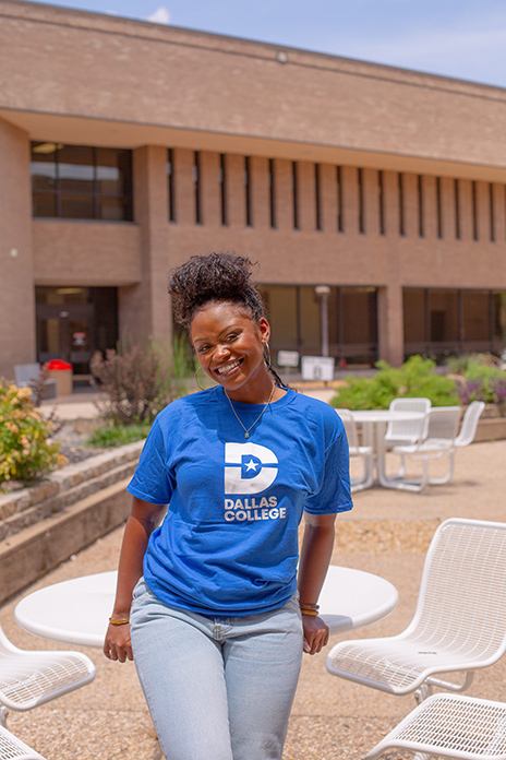 Kristen Bradford in a blue Dallas College shirt leans against a white, circular table.