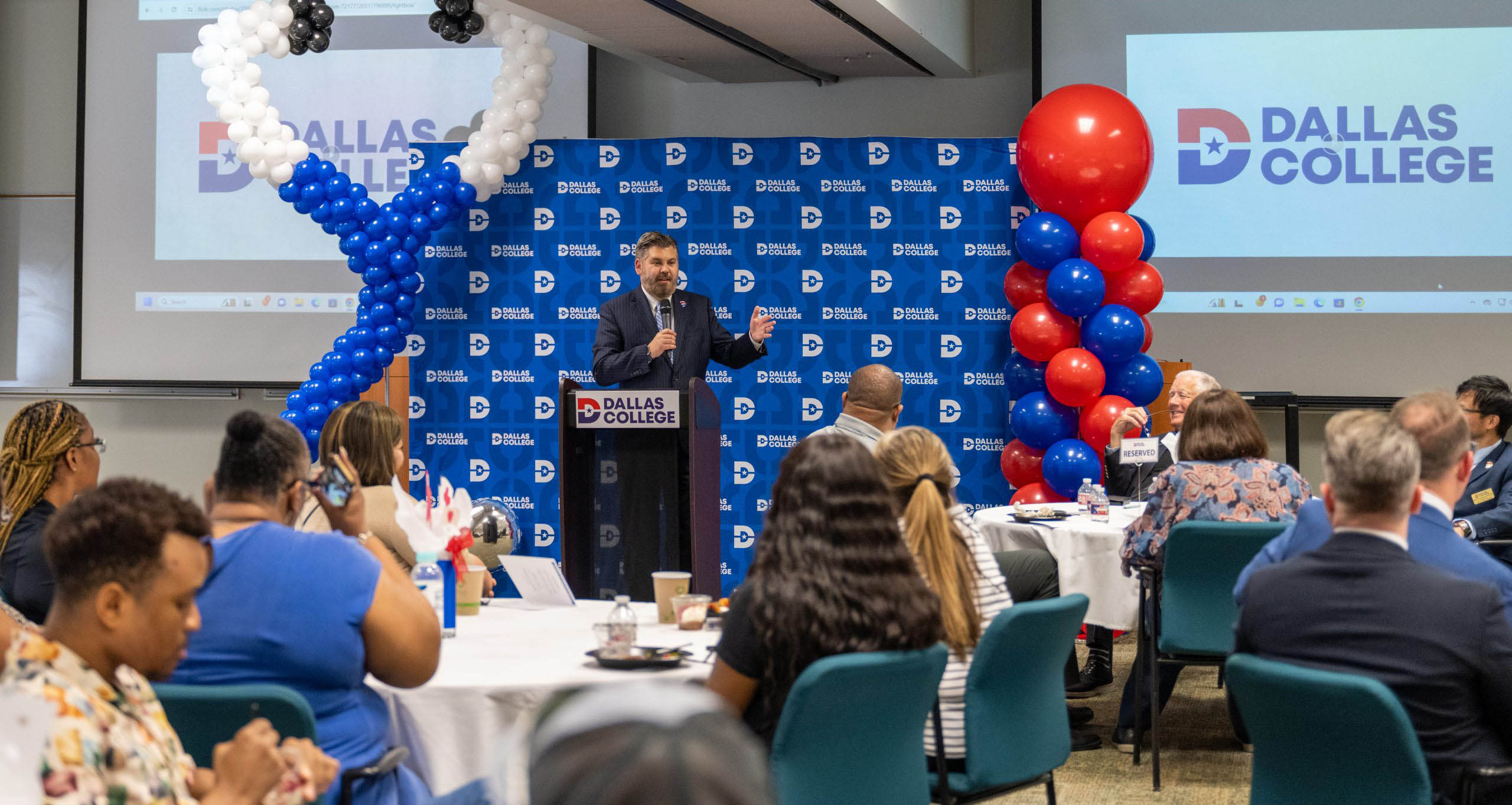 Lonon speaking at the RN-BSN Launch Party with a Dallas College backdrop behind him with tables filled with students with representatives and students listening.