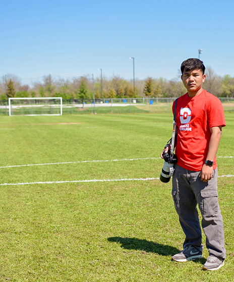 Ning wears a red Dallas College shirt with a camera strapped around his shoulder. He has the camera at his waist, with his hands on his side. As he faces the camera, the soccer field in the background is visible with goalposts setup.