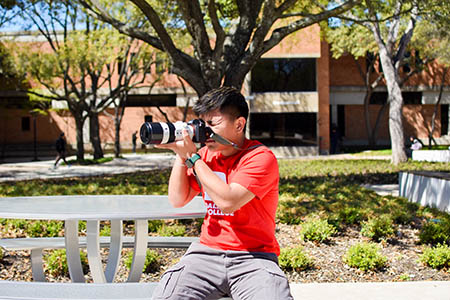Ning wearing a red Dallas College shirt with a camera strapped around his neck. He holds the camera at an eye-level with both hands, aiming the camera while sitting on a bench at a Dallas College campus.