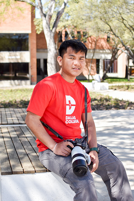 Ning wearing a red Dallas College shirt with a camera strapped around his neck. He holds the camera at a waist level, with one hand holding the camera and the other on his lap. He smiles at the camera while sitting on a bench at a Dallas College Campus.